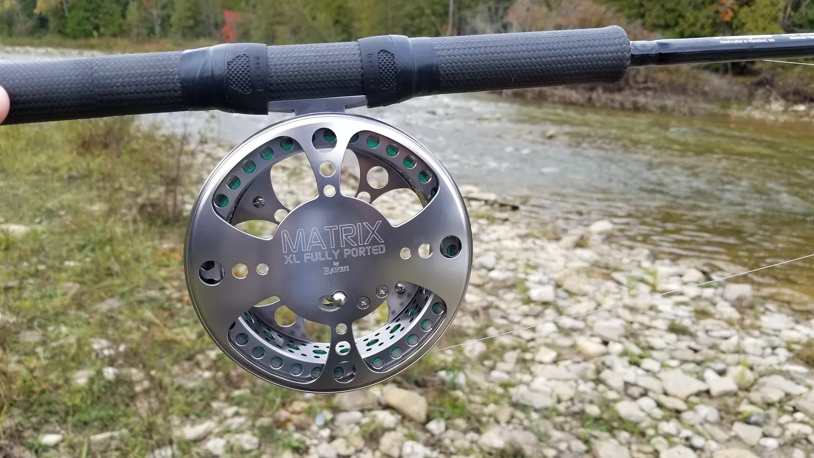 A centerpin reel on a long float rod beside a gravel river bank