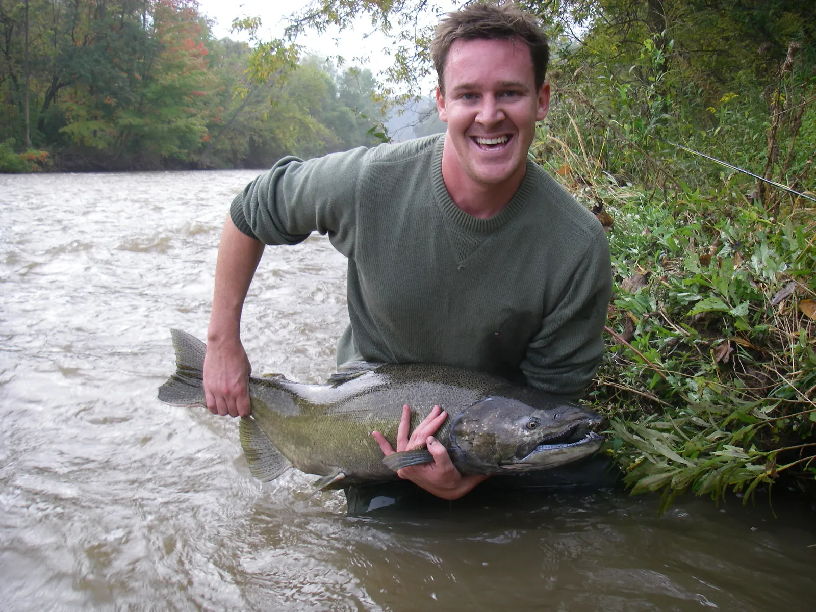 An angler crouched at the edge of the Lower Credit River with a chinook salmon
