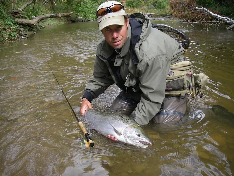 An angler kneeling in a Lake Ontario tributary with a chrome steelhead alongside a fly rod